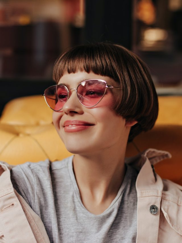 Cute girl with bright pink sunglasses sitting on yellow couch in cafe. Short-haired girl in light j